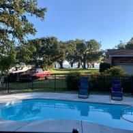 A backyard swimming pool with blue water sits in front of a metal fence, grassy lawn, mature trees, and a red truck.