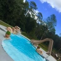 A bright blue swimming pool with a stone waterfall and a curved slide, surrounded by trees under a partly cloudy sky.