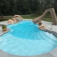 A curved backyard swimming pool with a rock water feature, tan slide, and a potted plant on the concrete patio.
