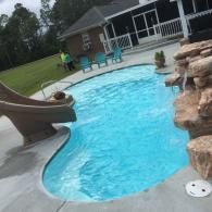 A swimming pool with a slide, a rock waterfall feature, and patio furniture in the backyard of a house.