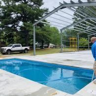 A person using a pool vacuum to clean an outdoor swimming pool near a partially constructed metal building and truck.