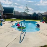 A backyard swimming pool with a turquoise interior, a pink unicorn pool float, and a black fence on a sunny day.