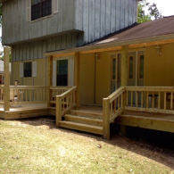 A newly built wooden deck with stairs and railings attached to a yellow house with a covered porch.