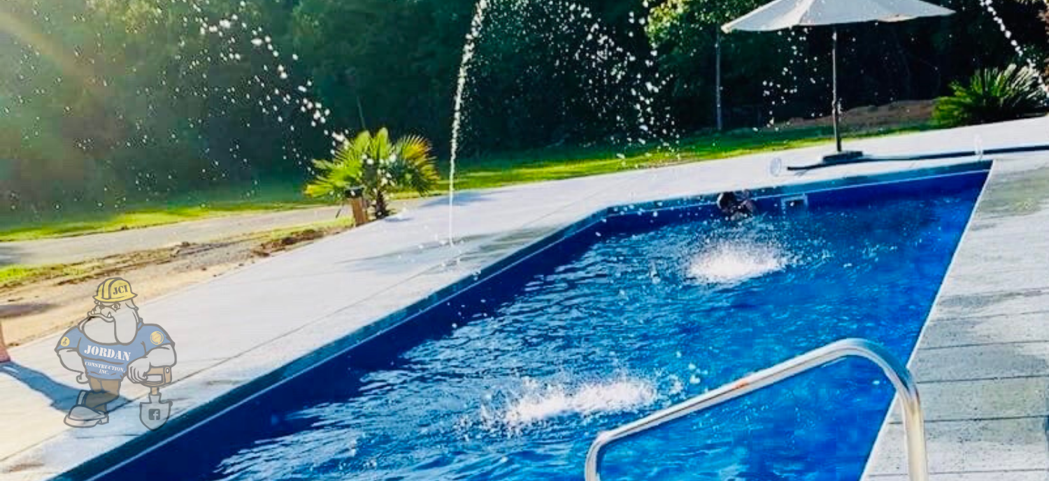 A blue rectangular swimming pool with decorative water fountains, poolside landscaping, and an umbrella on a sunny day.