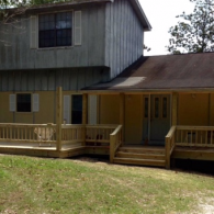 A two-story house with tan walls, gray vertical siding, and a newly built wooden wraparound deck and porch.