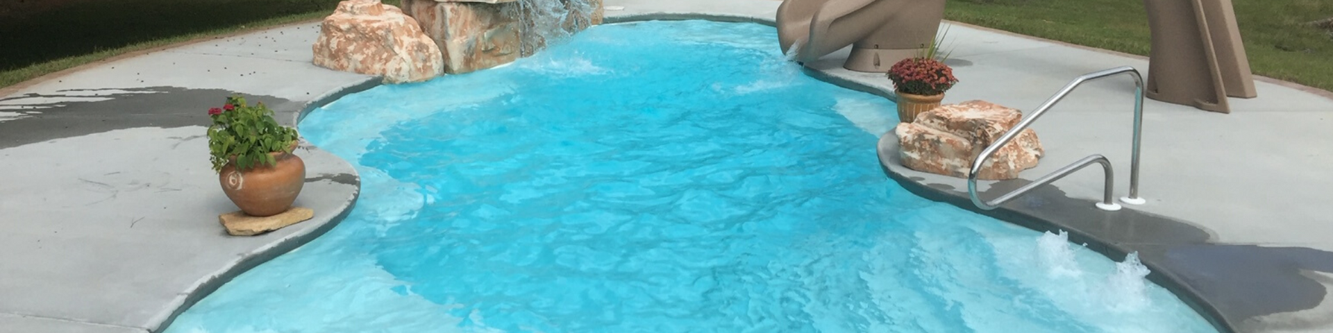 A blue backyard swimming pool featuring rock accents, a small slide, a ladder, and potted plants on the concrete deck.
