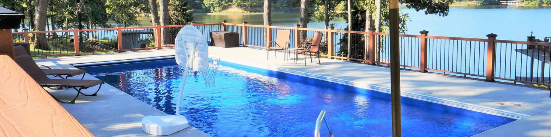 A rectangular blue pool on a patio deck overlooking a calm lake, surrounded by a wooden railing and outdoor lounge chairs.
