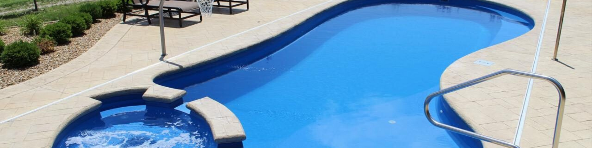 A blue kidney-shaped swimming pool with an integrated spa, surrounded by tan concrete decking and landscaping.