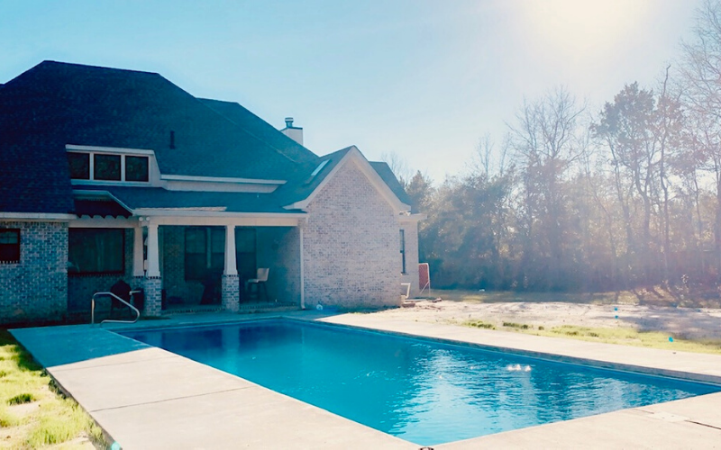 A rectangular swimming pool with bright blue water sits in the backyard of a multi-story brick house on a sunny day.