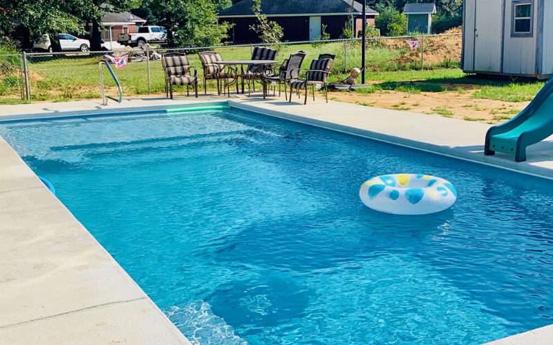 A sunny backyard swimming pool with a teal slide, a patterned floatie, and a patio set on the edge of the grass.