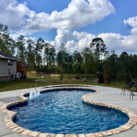 A kidney-shaped swimming pool with stone coping and a small water fountain sits in a backyard on a sunny, cloudy day.