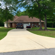 A brown brick house with a gabled roof, two cars parked in the garage driveway, surrounded by green trees and lawn.