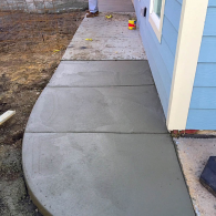 A freshly poured concrete sidewalk curves alongside a light blue house exterior with pale yellow trim.
