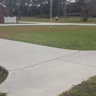 A concrete driveway curves through a green yard towards a white fence and trees under an overcast sky.