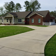 A single-story house with green siding and a red brick wing, featuring a covered front porch and a wide concrete driveway.