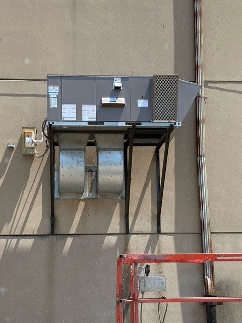 HVAC unit mounted on a building exterior. Gray metal, supported by a black frame; ventilation ducts are visible below.