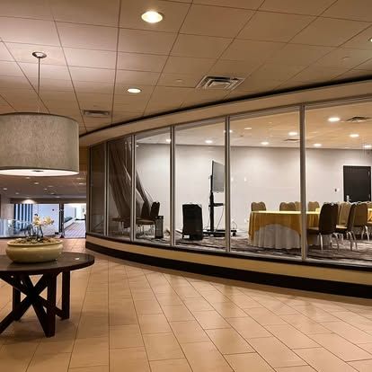 A hotel lobby with a conference room visible through large windows. A round table and pendant light are in the foreground.