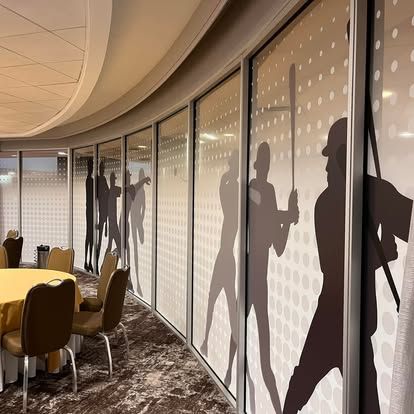 A curved room with frosted windows featuring baseball player silhouettes.  Tables with yellow tablecloths and chairs sit nearby.