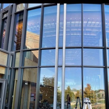 Modern building exterior with glass windows, metal framing, and reflective surfaces showing sky and surrounding buildings.