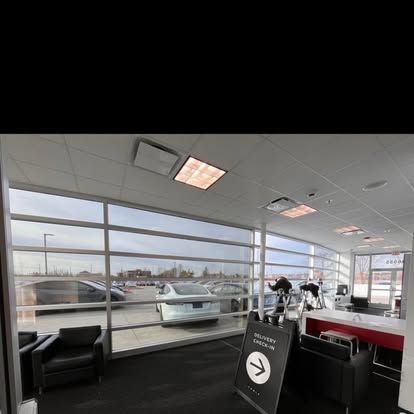 Interior view of a car dealership service area. Shows a delivery check-in sign, windows overlooking a parking lot, and black seating.