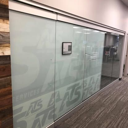 A glass-walled office with frosted logo lettering and a door, reflecting a workspace with chairs and a table.
