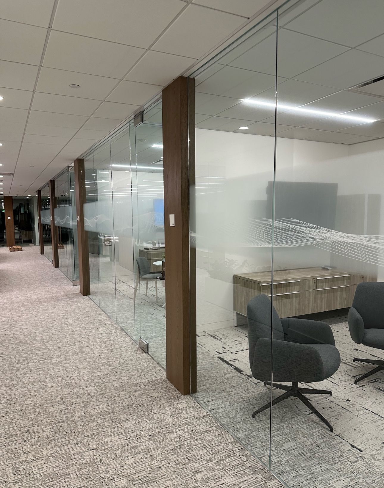 Office hallway with glass walls, a patterned carpet, and a seating area visible through the glass.