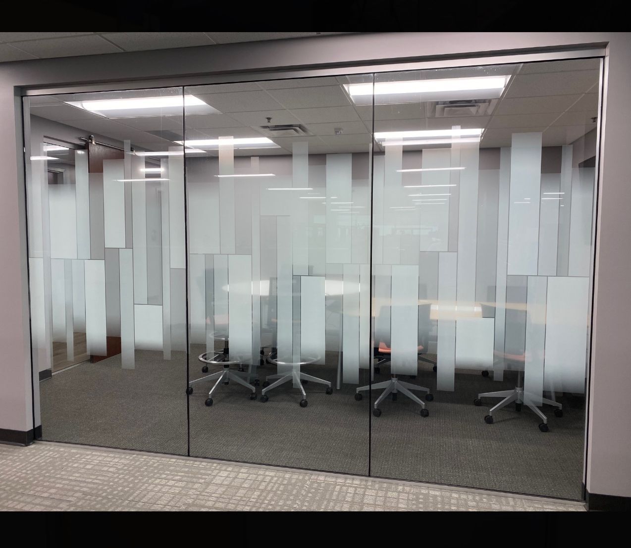 Glass-walled office with frosted rectangular patterns. Inside is a conference table with chairs, lit by overhead lights.