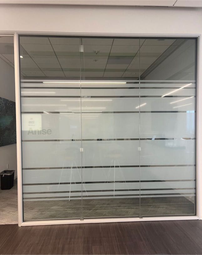 Glass office doors with frosted horizontal stripes and a logo. The doors are set in a white frame on a wooden floor.