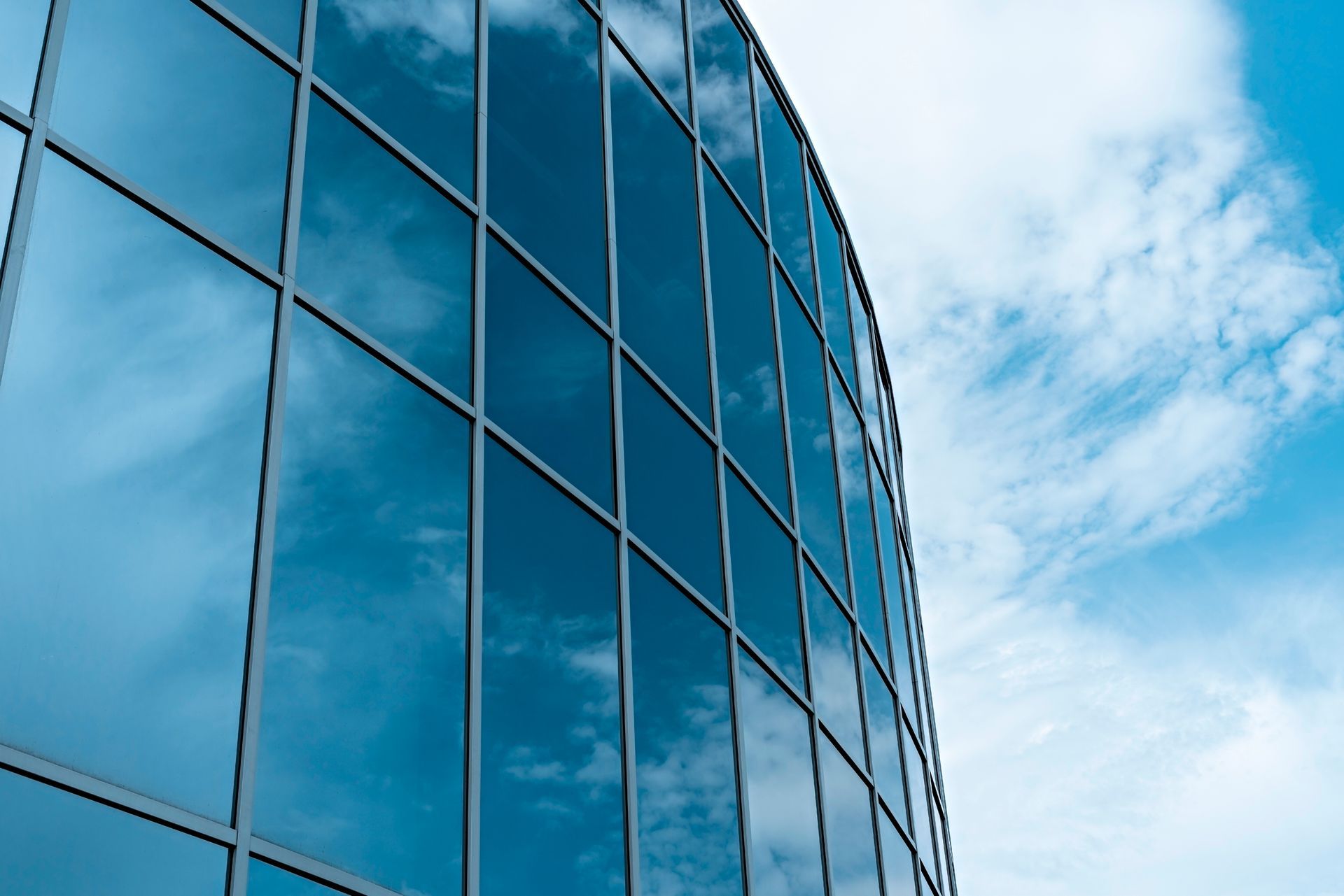 Glass facade of a modern building reflecting a bright blue sky with fluffy white clouds.