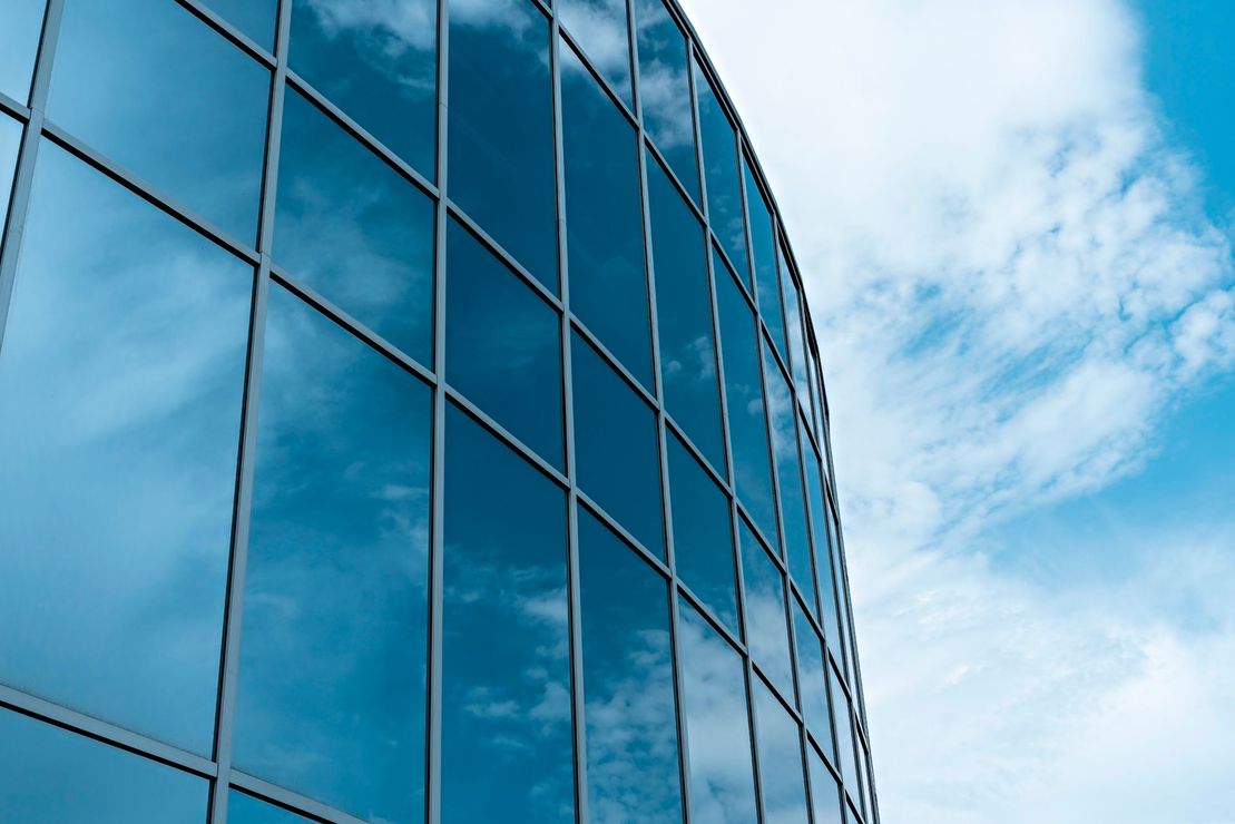 Curved glass facade of a modern building, reflecting a bright blue sky with white clouds.