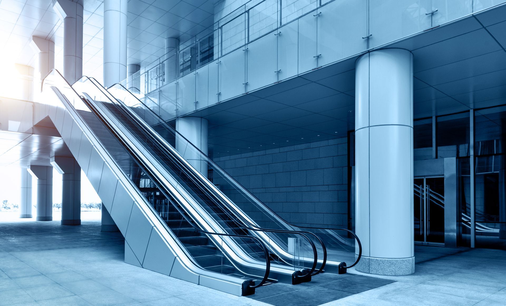Escalators ascending inside a modern building. Blue-toned lighting and architecture with large pillars.