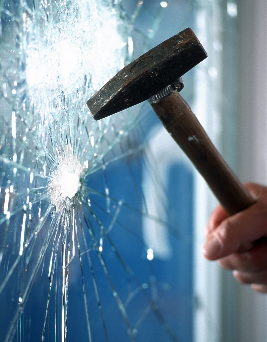 A hand swings a hammer, smashing a window; shards of glass radiate from the impact point.