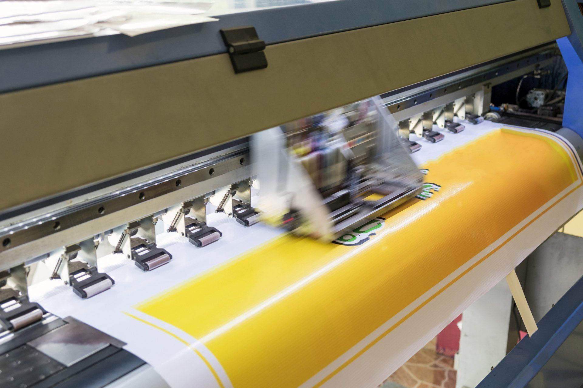 Large format printer in operation, spraying ink on yellow and white poster paper. The machine is beige and blue.