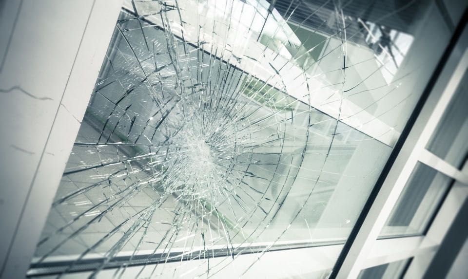 Shattered window with cracks radiating from the center. Light and blurred greenery are visible through the broken glass.