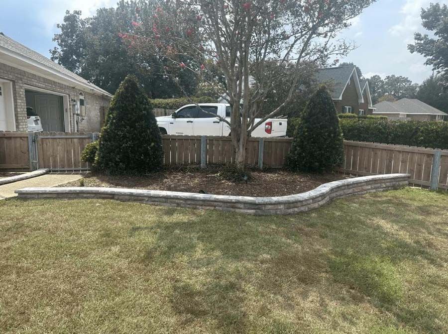 Lawn with a brick wall around a tree and two bushes, a white truck, a house, and fence.