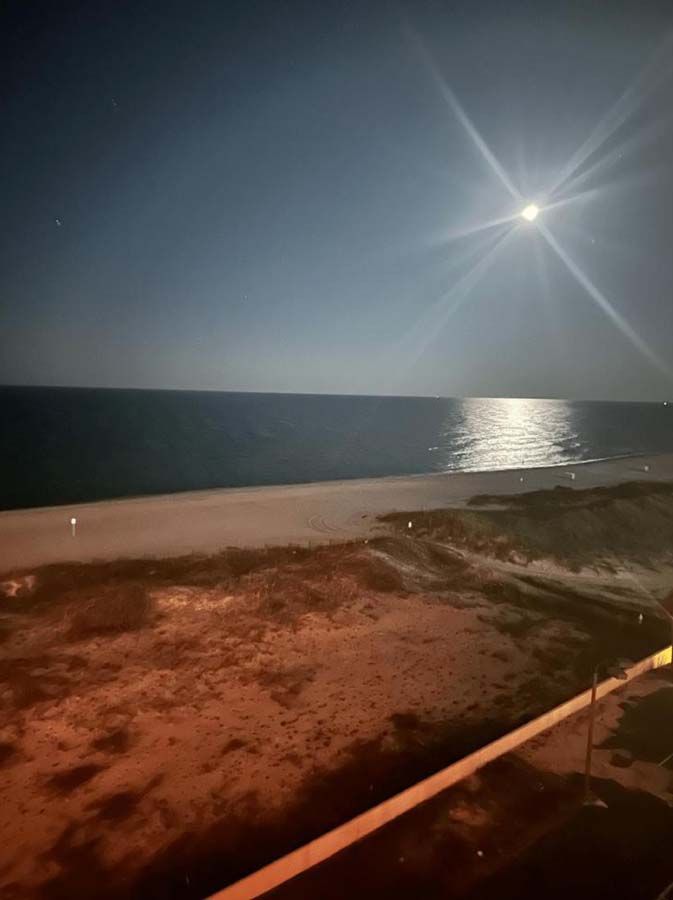 Moonlit beach scene at night. Moon reflected on water, dark sky, orange glow on foreground.