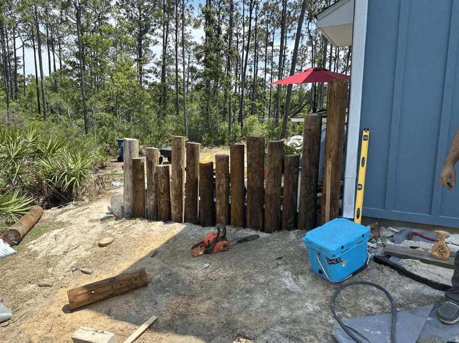 Wooden fence under construction next to a blue building, with a chainsaw and cooler nearby.