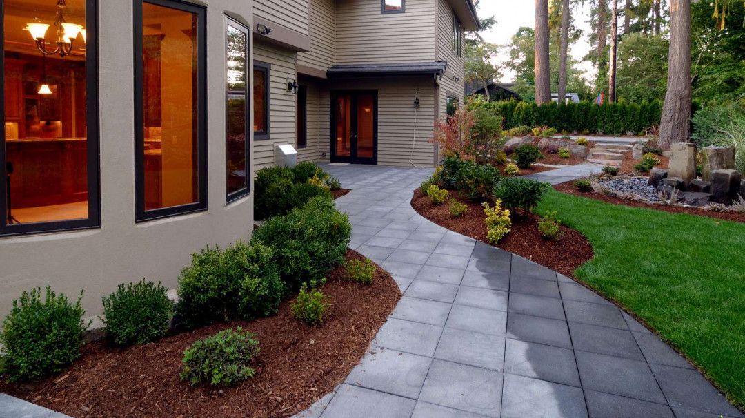 Paver pathway curving from house with landscaping, surrounded by green lawn and trees.