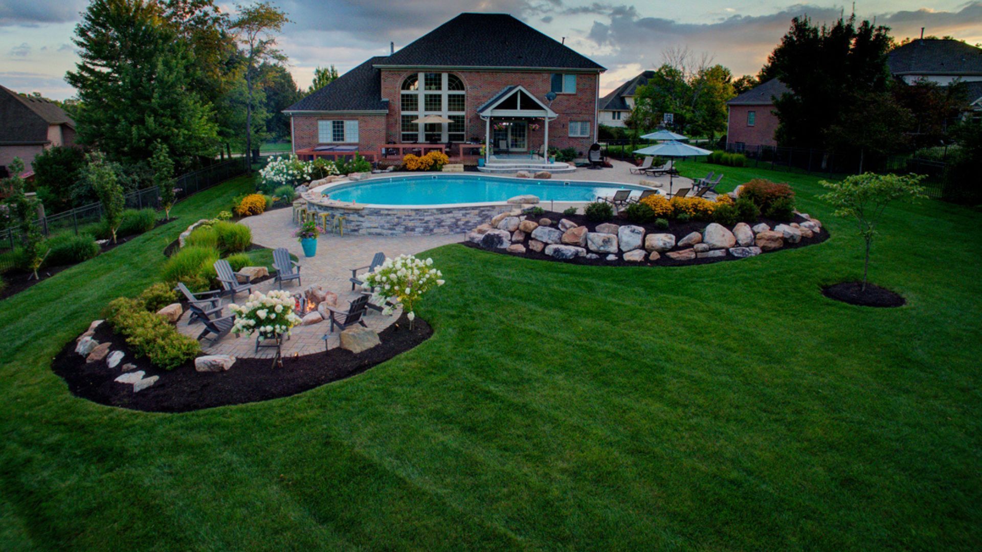 Backyard with pool, landscaping, and large brick house at sunset.