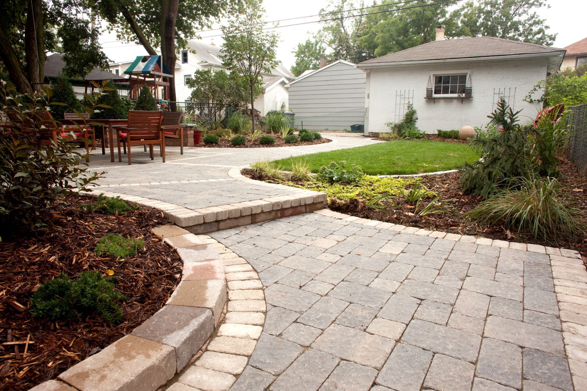 Brick pathway in backyard leading to a patio and a garage. Landscaping and grass border the path.