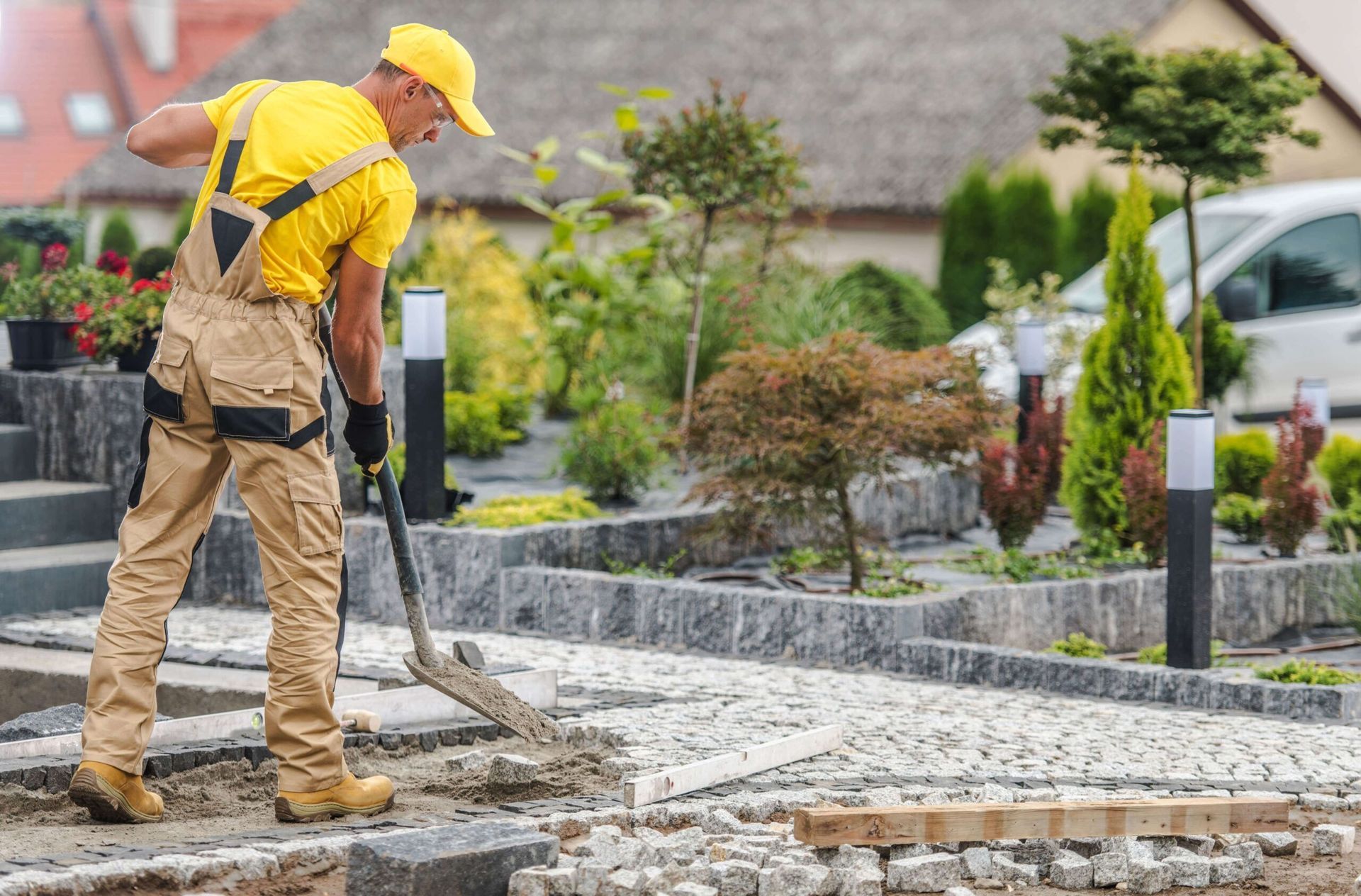 Man in yellow shirt and overalls shoveling soil on a stone pathway in a garden.