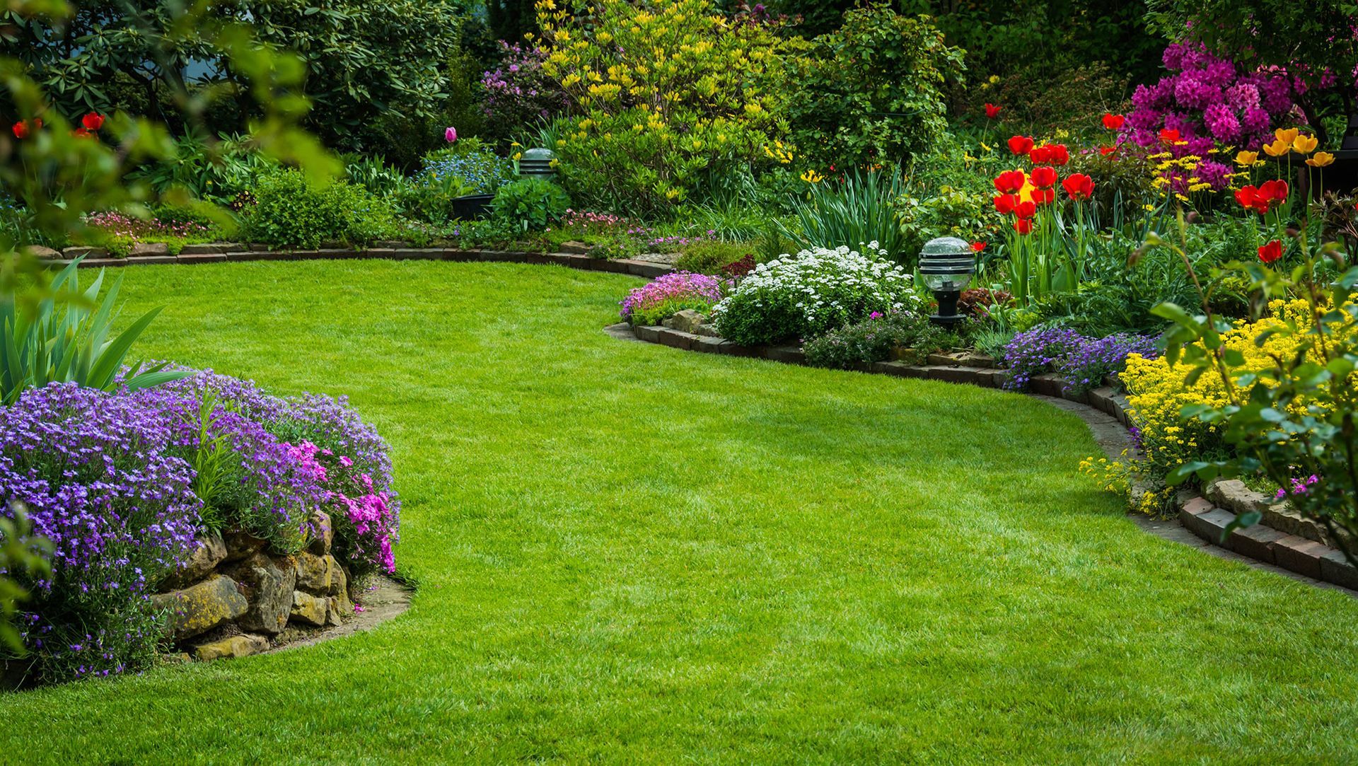 Lush green lawn with colorful flower beds, bordered by stone, in a sunny garden.
