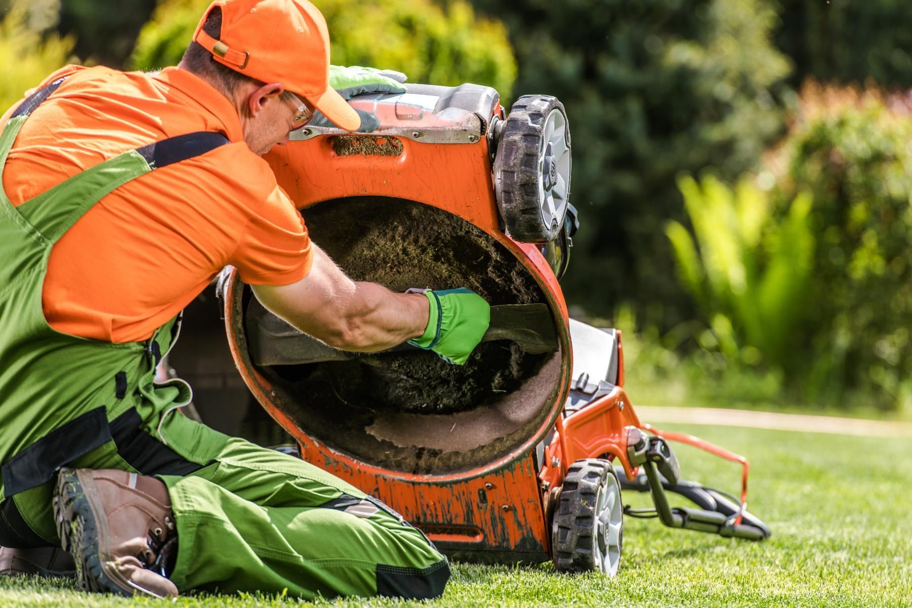Person in orange outfit cleaning a lawnmower on a green lawn.