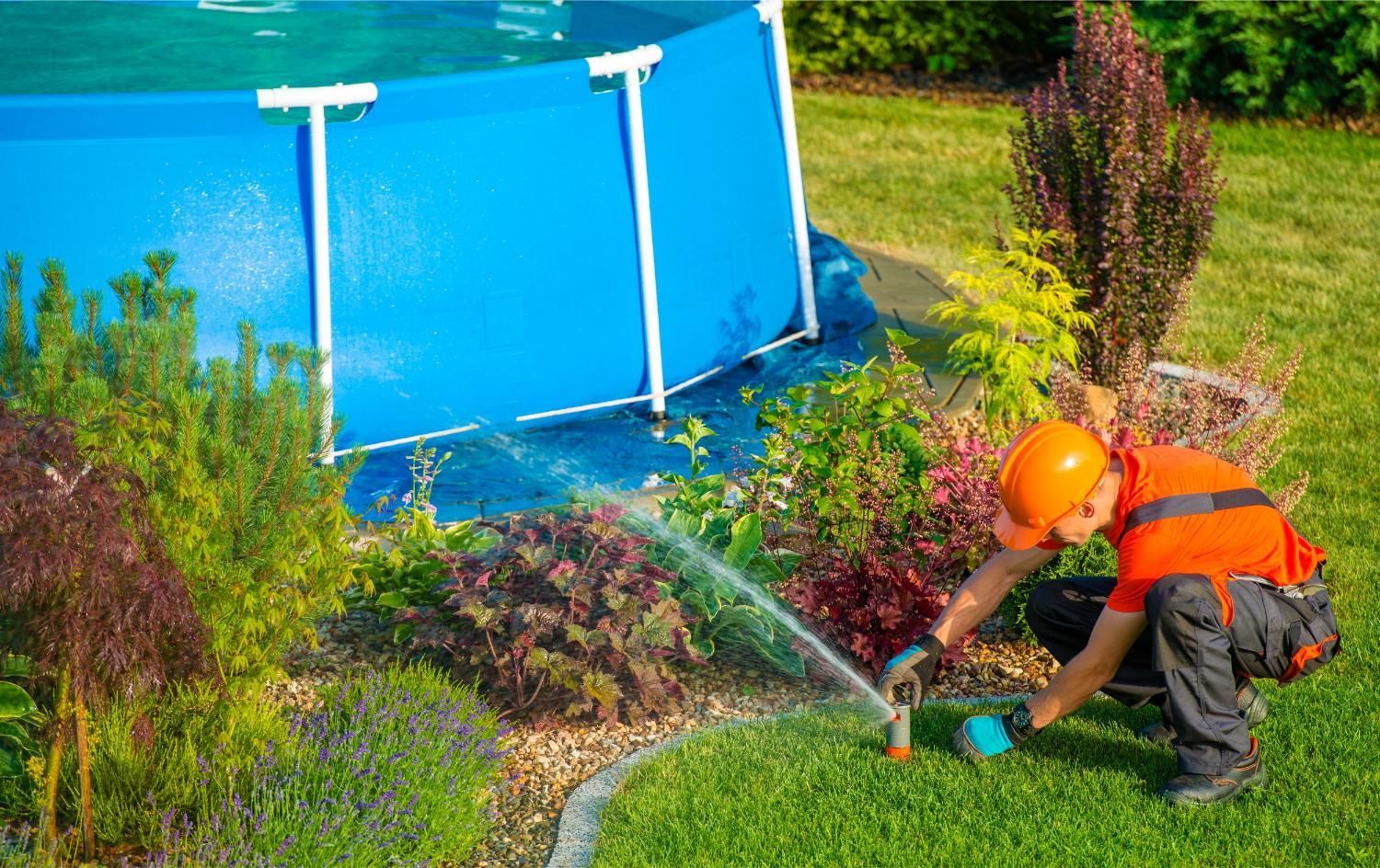 Man in orange gear installing sprinkler near a blue pool and garden.