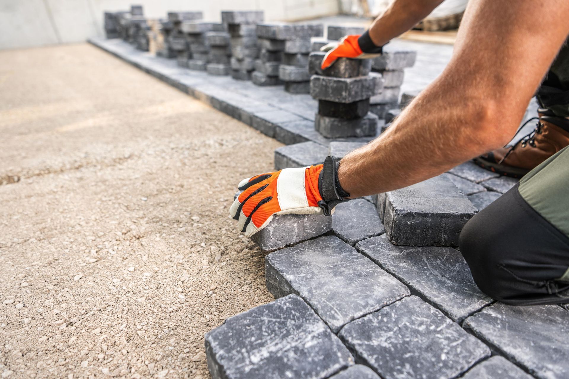 Person wearing work gloves laying dark pavers on a gravel bed.