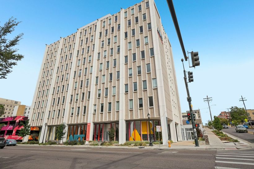 Multi-story building with vertical panels, windows, and colorful ground-level storefronts at a street intersection.