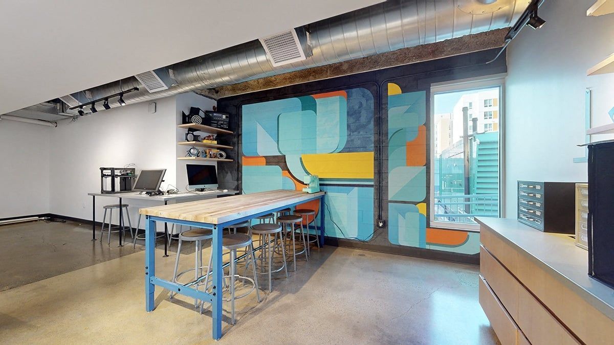 Interior with a long blue table, colorful mural, concrete floor, and shelves with equipment.