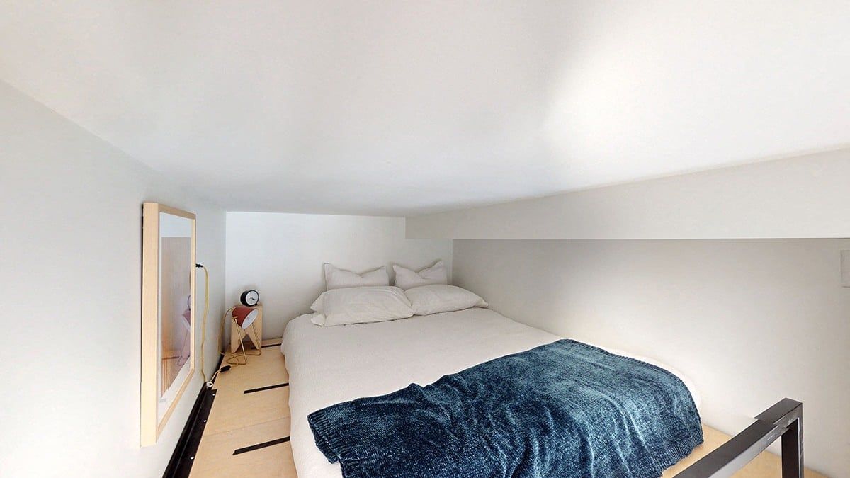 Small bedroom with bed, pillows, and a blue blanket. Mirror on the wall.