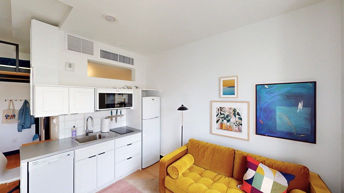 Small, modern apartment interior with yellow sofa, white kitchen, and artwork on the wall.