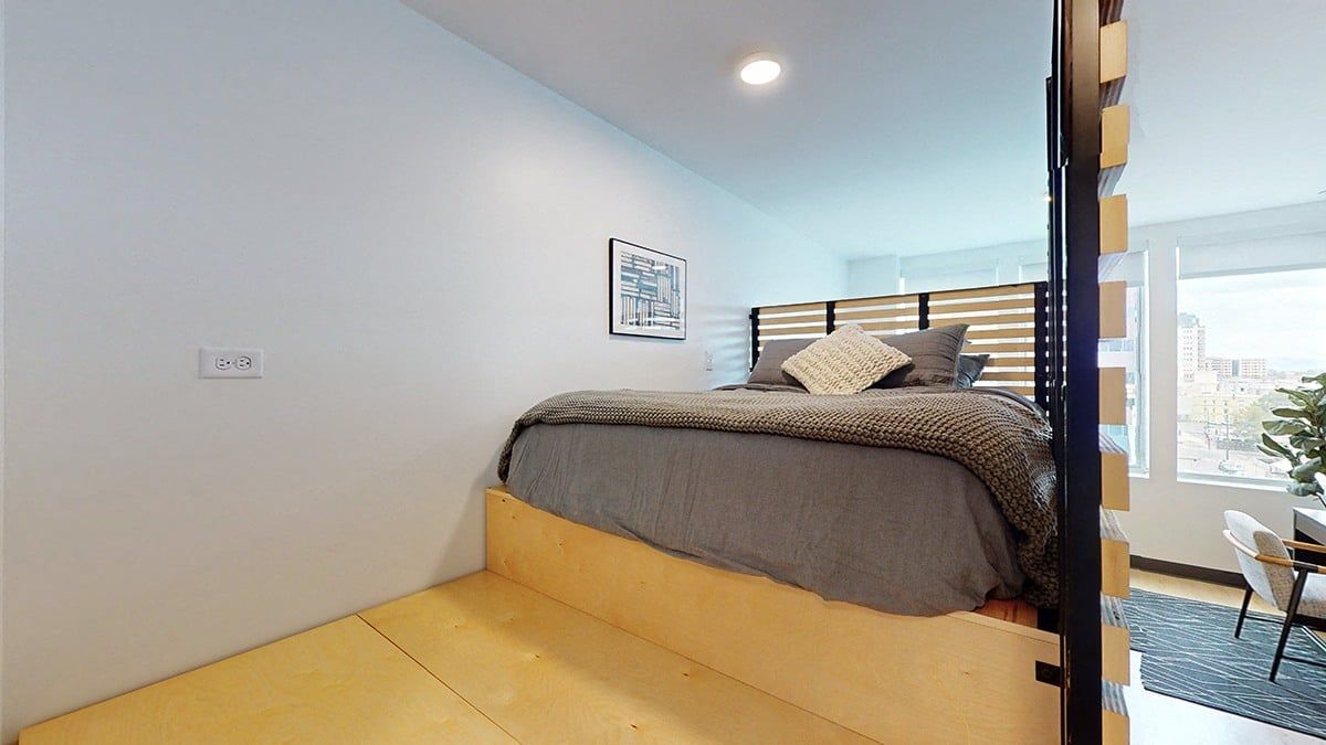 Loft bedroom with elevated bed, light wood platform, gray bedding, neutral walls, and window.